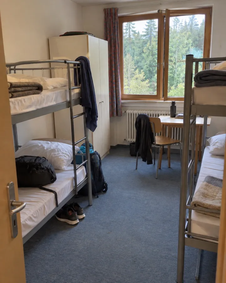 Blick von der Tür in ein schlichtes Mehrbettzimmer mit zwei Etagenbetten, einem Schrank und einem Tisch am Fenster zum Wald; auf einem Bett liegt ein Rucksack.