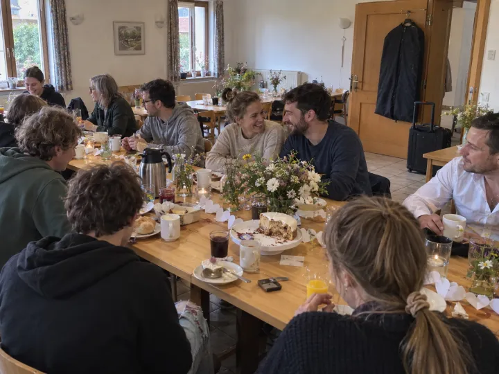 Spätes Frühstück nach der Feier: Gäste in Alltagskleidung sitzen an langen Holztischen mit Wiesenblumen, Kaffee und einem angeschnittenen Kuchen, in der Mitte lacht ein Paar.