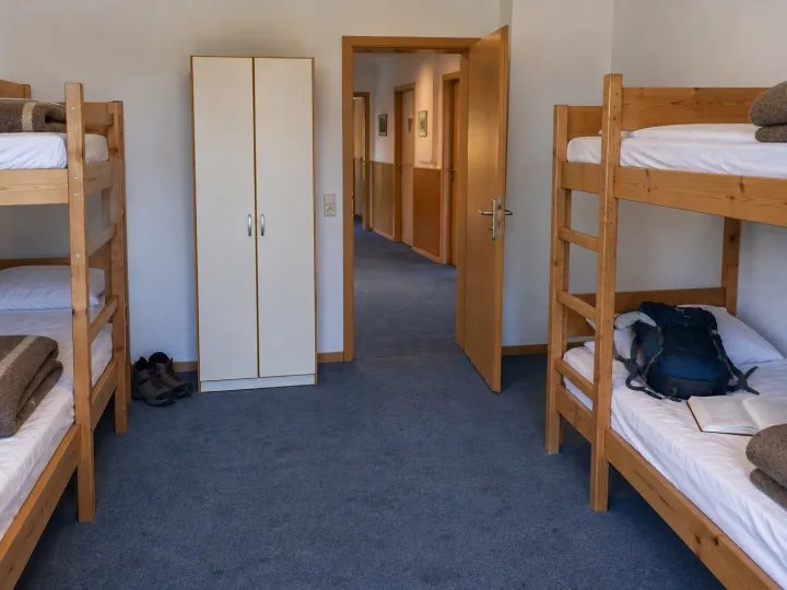 Schlichtes Zimmer mit zwei Etagenbetten aus Holz und einem hellen Schrank; die Tür steht offen zum Flur, auf einem Bett liegen ein Rucksack und ein Buch.
