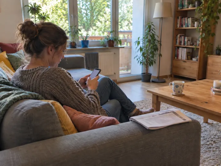 Eine Frau sitzt auf dem Sofa im Wohnzimmer und tippt auf ihrem Handy, von schräg hinten gesehen; auf der Armlehne liegt ein gefaltetes Blatt.